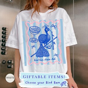 May include: White t-shirt with a blue and pink graphic. The graphic features a peacock, cocktail glass, and bamboo. Text includes "bird ban cheers club" and "GIFTABLE ITEMS! Choose your Bird Bam".
