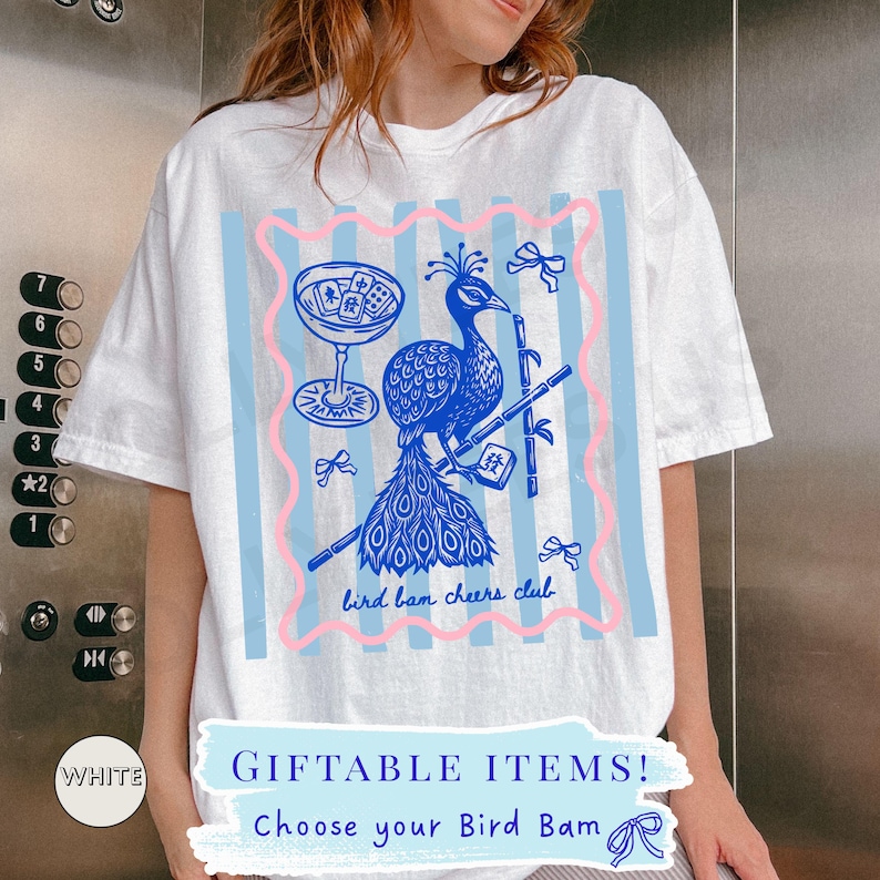 May include: White t-shirt with a blue and pink graphic. The graphic features a peacock, cocktail glass, and bamboo. Text includes "bird ban cheers club" and "GIFTABLE ITEMS! Choose your Bird Bam".