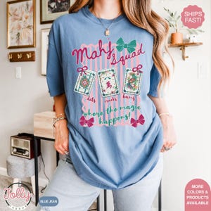 May include: A blue jean colored t-shirt with the text "Mahj Squad" in pink script. Below the text are three Mahjong tiles and the words "dots, cracks, bams, where the magic happens!" in a pink and teal design.
