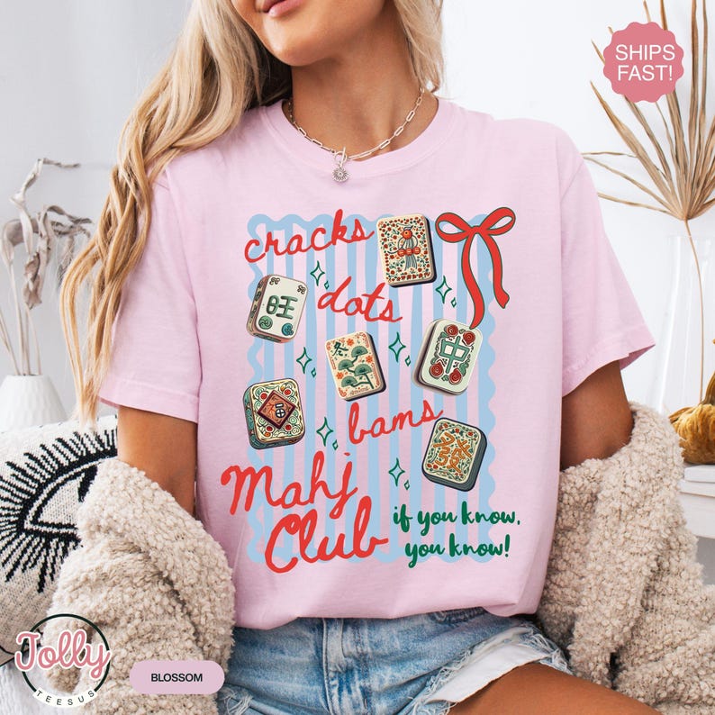 May include: A light pink t-shirt with a graphic design featuring Mahjong tiles, the words "cracks, dots, bams, Mahj Club" and the phrase "if you know, you know!" in red and blue. The shirt has a relaxed fit.
