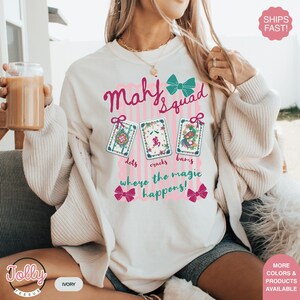 May include: Ivory-colored sweatshirt with the text "Mahj Squad" in pink script and a teal bow. Below are three illustrated Mahjong tiles and the phrase "where the magic happens!" in pink. The Jolly Teesub logo is in the bottom left.