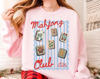 May include: Light pink sweatshirt with a graphic of Mahjong tiles and the words "Mahjong Club" in red script. The design includes blue vertical stripes and the phrase "all day everyday".