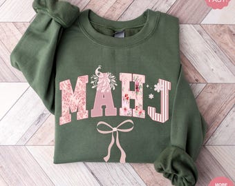 May include: Military green sweatshirt with the letters "MAHJ" in a floral and striped design. A pink bow is below the letters. The sleeves are cuffed. The text "SHIPS FAST!" and "MORE COLORS & PRODUCTS AVAILABLE" are in the image.