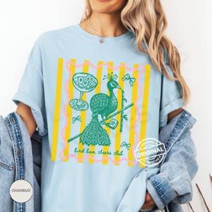 May include: Light blue t-shirt with a graphic of a green peacock, cocktail glass, and bamboo. The design is framed by yellow stripes and pink wavy lines. Text reads "bird bam cheers club."