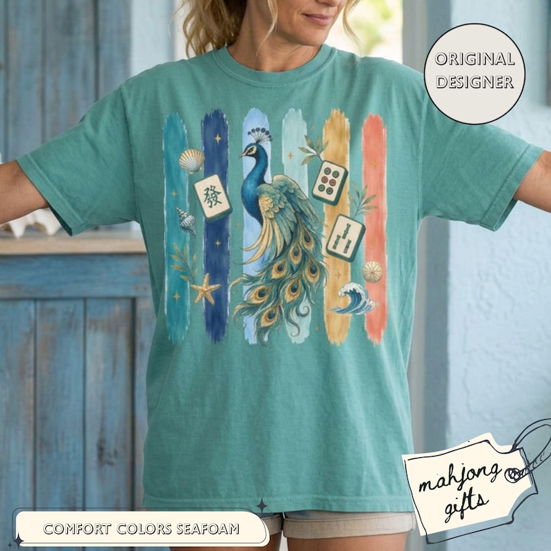 May include: Seafoam green t-shirt with a peacock design and colourful vertical stripes. The peacock has blue and gold feathers, and the shirt includes Mahjong tiles, seashells, and the text "Comfort Colors Seafoam".