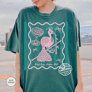 May include: Teal-colored t-shirt featuring a pink peacock design within a wavy-edged frame. The design includes a cocktail glass, bamboo, and the text "bird ban cheers club." A "ORIGINAL" stamp is visible.