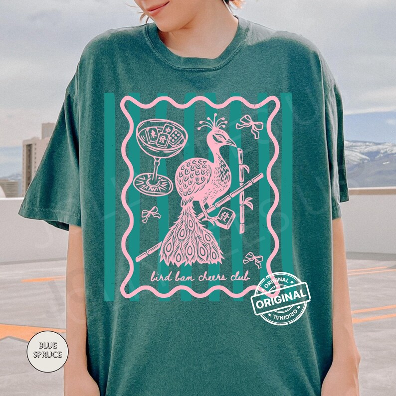 May include: Teal-colored t-shirt featuring a pink peacock design within a wavy-edged frame. The design includes a cocktail glass, bamboo, and the text "bird ban cheers club." A "ORIGINAL" stamp is visible.