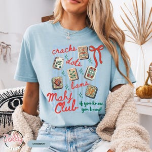 May include: Light blue t-shirt with a graphic of Mahjong tiles and the words "cracks, dots, bams, Mahj Club, if you know, you know!" in red and blue. The shirt is made of a soft material.