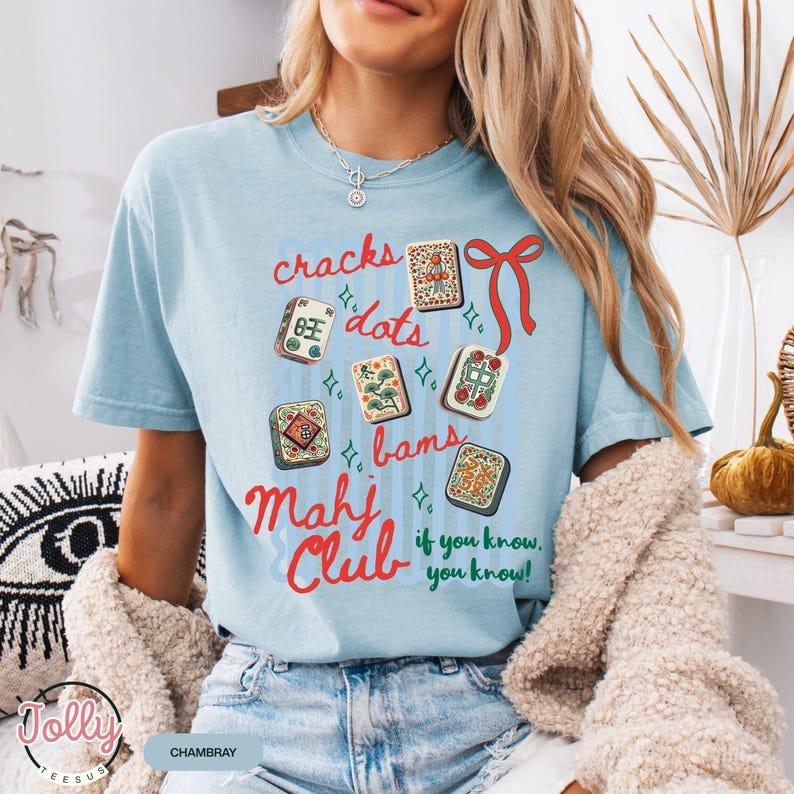 May include: Light blue t-shirt with a graphic of Mahjong tiles and the words "cracks, dots, bams, Mahj Club, if you know, you know!" in red and blue. The shirt is made of a soft material.