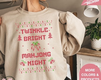 May include: Beige sweatshirt with a festive design. The text reads "Twinkle Bright Mahjong Night" in pink, with Christmas tree and bell graphics. The design is reminiscent of a holiday sweater.