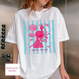 May include: White t-shirt featuring a pink peacock design with a cocktail glass and bamboo. The design includes the text "bird ban cheers club" and is set against a blue striped background. The shirt also has the text "Shocking Pink Bird Bam".
