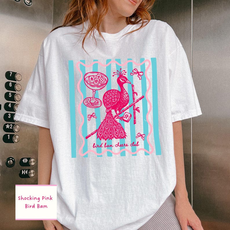 May include: White t-shirt featuring a pink peacock design with a cocktail glass and bamboo. The design includes the text "bird ban cheers club" and is set against a blue striped background. The shirt also has the text "Shocking Pink Bird Bam".