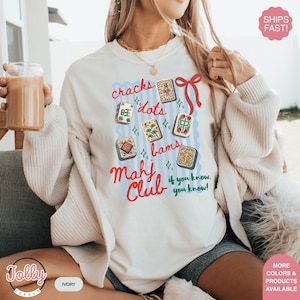 May include: Ivory-colored sweatshirt featuring a colorful Mahjong-themed design. The design includes the words "cracks, dots, bams" and "Mahj Club" with various Mahjong tiles. The text "if you know, you know!" is also included.