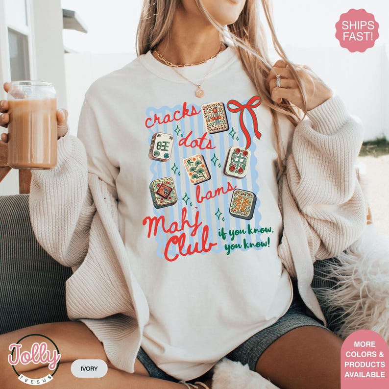 May include: Ivory-colored sweatshirt featuring a colorful Mahjong-themed design. The design includes the words "cracks, dots, bams" and "Mahj Club" with various Mahjong tiles. The text "if you know, you know!" is also included.