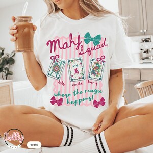 May include: White t-shirt with a pink and white striped design. The shirt features the text "Mahj Squad" in pink script, along with illustrations of Mahjong tiles and the phrase "where the magic happens!" in a playful font.