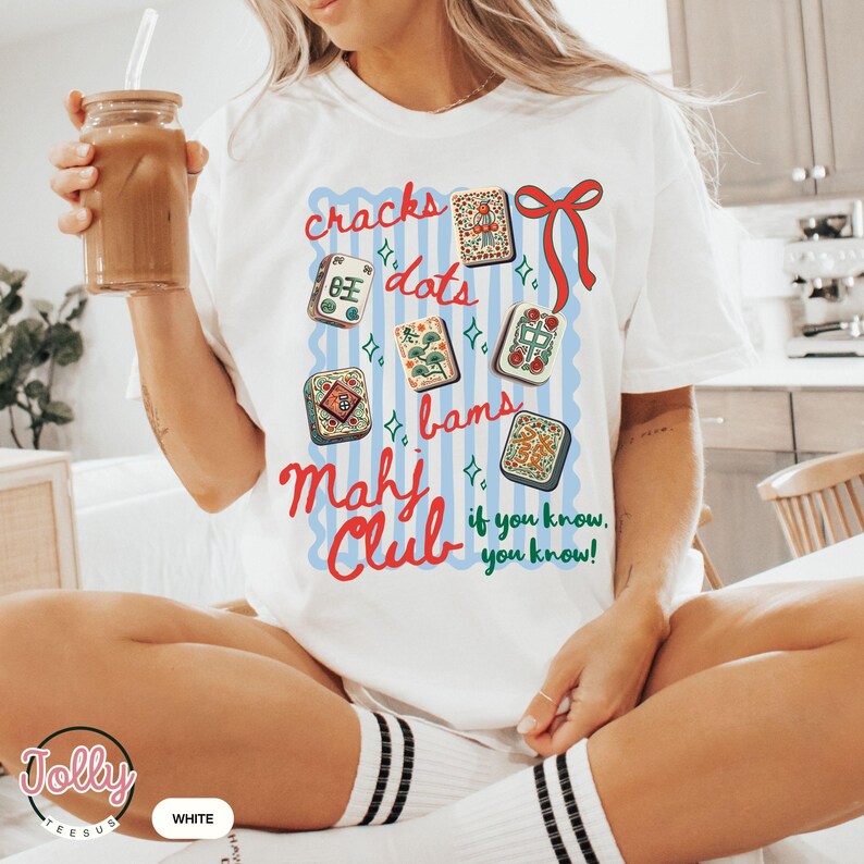 May include: White t-shirt with a colorful graphic design featuring Mahjong tiles and the words "cracks, dots, bams, Mahj Club, if you know, you know!" The shirt has a crew neck and short sleeves.