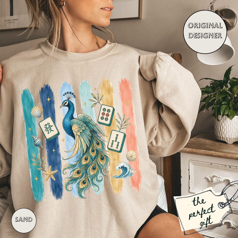 May include: A sand-coloured sweatshirt with a peacock design and colourful brushstrokes. The peacock is in shades of blue and green, with Mahjong tiles and other decorative elements. The text "Original Designer" is in a circle.
