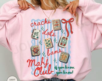 May include: A light pink crewneck sweatshirt featuring a graphic print of Mahjong tiles, a red ribbon, and blue vertical stripes. The text reads: cracks, dots, bams, Mahj Club, if you know, you know! The design includes small green star accents.