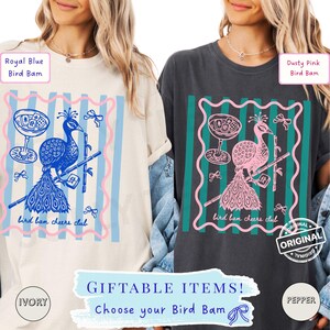 May include: Two t-shirts, one ivory and one pepper, featuring a peacock design with the text "bird bam cheers club." The design includes a cocktail glass and bamboo. The text "GIFTABLE ITEMS! Choose your Bird Bam" is also present.