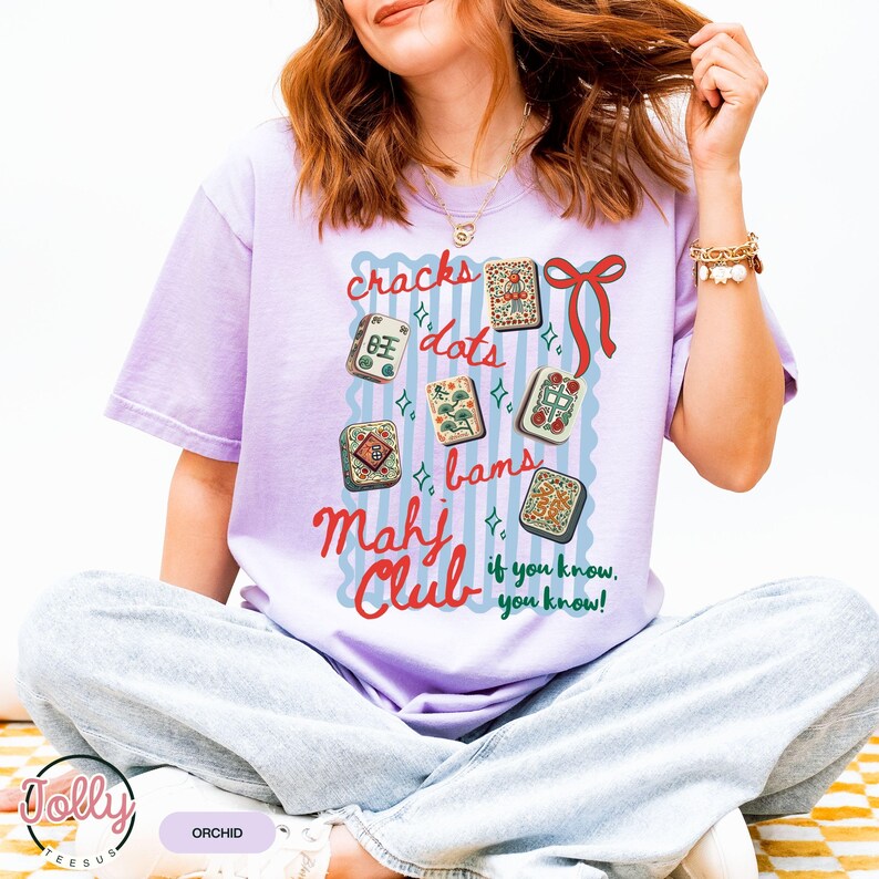 May include: Orchid-colored t-shirt with a graphic design featuring Mahjong tiles and the words "cracks, dots, bams, Mahj Club, if you know, you know!" The design includes a red bow and blue accents. The shirt is a relaxed fit.