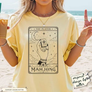 Peut inclure: T-shirt jaune beurre avec un motif de carte de tarot. Le graphisme présente les mots "THE PLAYER" et "MAHJONG", une main tenant une tuile avec des caractères chinois et des éléments célestes. Le t-shirt a une coupe décontractée.