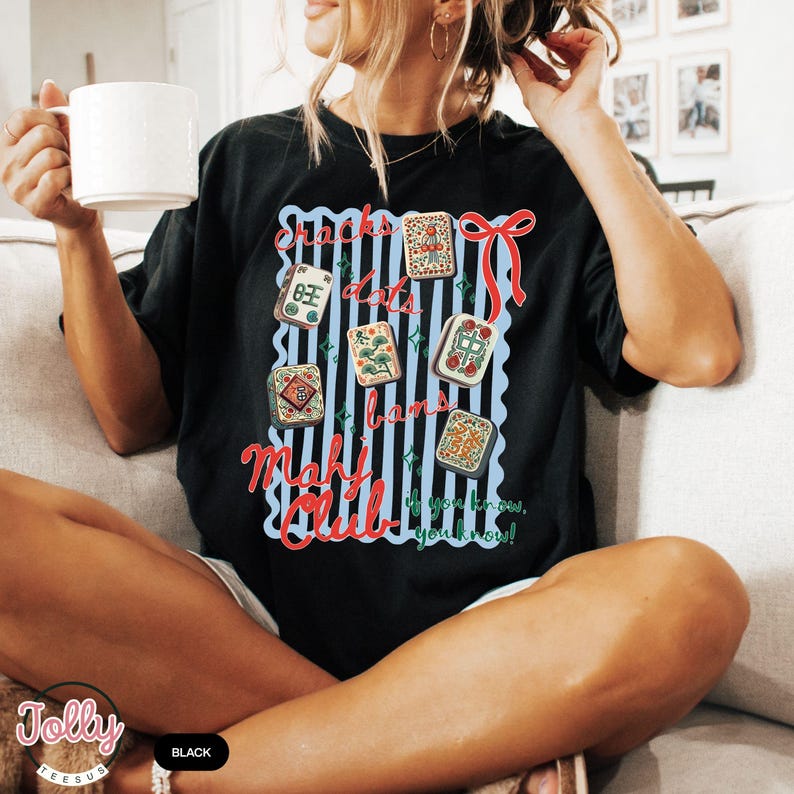 May include: Black t-shirt with a colorful graphic featuring Mahjong tiles, a red bow, and the words "Cracks, Dots, Bams, Mahj Club." The design is set against a blue and white striped background. The shirt also has the text "If you know, you know!"