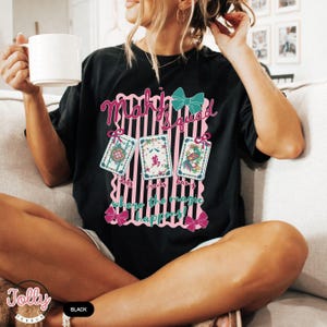 May include: Black t-shirt with a pink and teal graphic that reads "Mahj Squad" and "dots cracks bangs where the magic happens!" The graphic includes three illustrated cards and bows. The shirt is a relaxed fit.