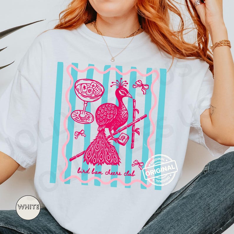 May include: White t-shirt with a pink and blue graphic. The graphic features a pink peacock with bamboo, a cocktail glass, and the text "bird bam cheers club." The design is framed by pink lines and blue stripes.
