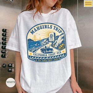 Peut inclure: T-shirt blanc avec un motif bleu et jaune. Le motif indique "MAHGIRLS TRIP CALIFORNIA COAST PCH EST. GIRLS ONLY 2026" et présente une scène côtière avec une voiture, un pont et le soleil.