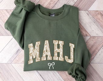 May include: An olive green sweatshirt with the word "MAHJ" in a floral design across the chest, with a small white bow underneath. The sweatshirt is folded on a light brown wooden surface.