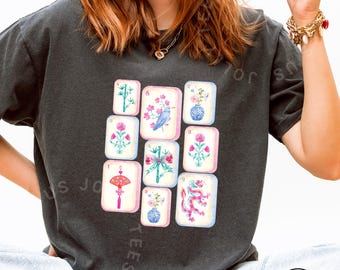 May include: Dark gray t-shirt featuring a watercolor design of nine Mahjong tiles. The tiles display various floral and bird illustrations in pink, blue, and green. The word "Pepper" is in a white circle at the bottom right.