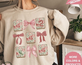 May include: Beige sweatshirt featuring a design of pink bows and floral tiles with Chinese characters. The design includes a dragon, flowers, and the word "Jolly" at the bottom left. The sweatshirt has a crew neck.
