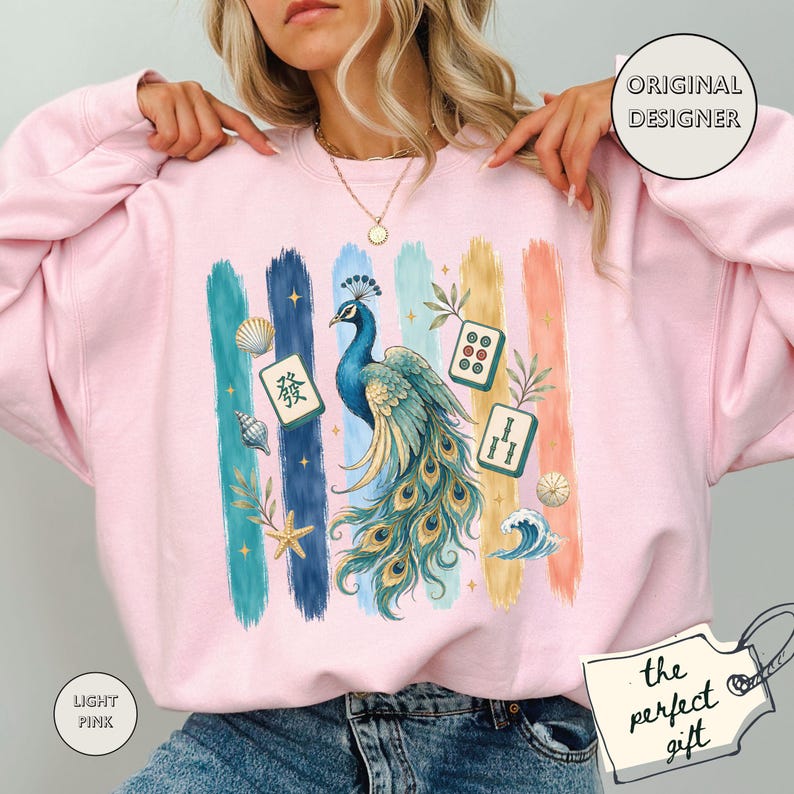 May include: Pale pink sweatshirt featuring a peacock design and vertical stripes in various colours. The peacock is blue and teal with a long tail. The design includes seashells, starfish, and Mahjong tiles. The text "ORIGINAL DESIGNER" is in a circle.