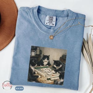May include: Blue short-sleeve t-shirt with a graphic of three black and white cats playing a game. The shirt has a crew neck and the text "Comfort Colors" on the tag. A gold necklace is draped across the shirt.