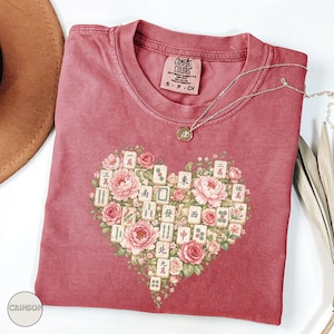 May include: Crimson-colored t-shirt with a heart-shaped design of pink flowers and Mahjong tiles. The shirt has a crew neck and short sleeves. The brand label "COMFORT COLORS" is visible on the neckline. A silver necklace with a pendant is draped on the shirt.