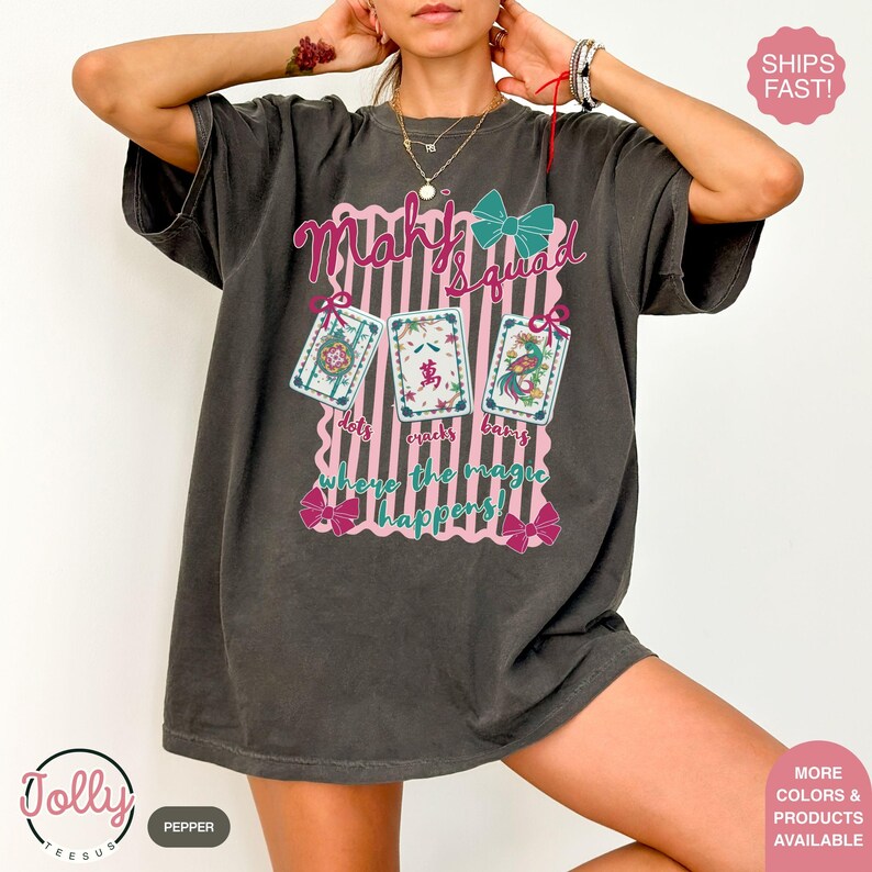 May include: Dark grey t-shirt with the text "Mahy Squad" and three illustrated cards. The design includes pink vertical stripes, bows, and the phrase "dots cracks bangs where the magic happens!"
