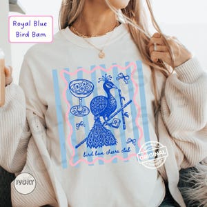May include: Ivory-colored sweatshirt featuring a royal blue peacock design with a cocktail glass and bamboo. The design is framed with pink and blue stripes, and the text "bird bam cheers club" is visible. The words "Royal Blue Bird Bam" are in a box at the top.