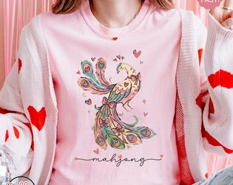 May include: A light pink crew neck t-shirt featuring a colorful peacock design with hearts and bows. The word "mahjong" is written in cursive below the peacock. The shirt is part of the "Blossom" collection.