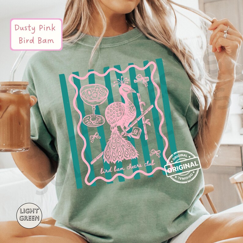 May include: Light green t-shirt with a pink peacock graphic inside a teal frame. The text "Dusty Pink Bird Bam" is at the top left, and "bird bam cheers club" is at the bottom. The shirt has a relaxed fit.
