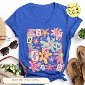May include: A heather true royal blue V-neck t-shirt featuring a colorful graphic print of various flowers and Mahjong tiles. The design includes multiple floral shapes in pink, yellow, and light blue, interspersed with Mahjong tiles.