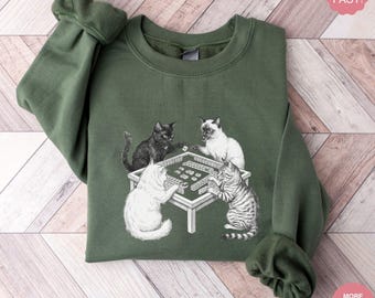 Peut inclure: Sweat-shirt vert militaire avec un motif de quatre chats jouant au mahjong. Les chats sont noirs, blancs et gris, et la table de jeu est blanche. Le sweat-shirt est de couleur unie avec des poignets côtelés.