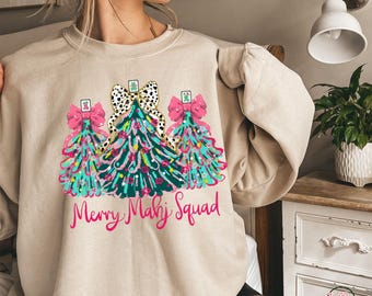 May include: Beige sweatshirt featuring a colorful graphic of three Christmas trees with pink bows and a black and white polka dot bow. The text "Merry Mahj Squad" is written in pink script below the trees. The sweatshirt is a casual, festive garment.