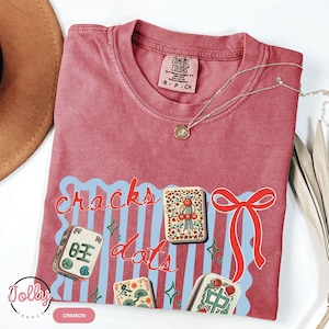 May include: Crimson-colored t-shirt with the words "Cracks Dots" in red script over a light blue wavy design. The shirt features illustrations of Mahjong tiles and a red bow. A gold necklace is draped across the shirt.
