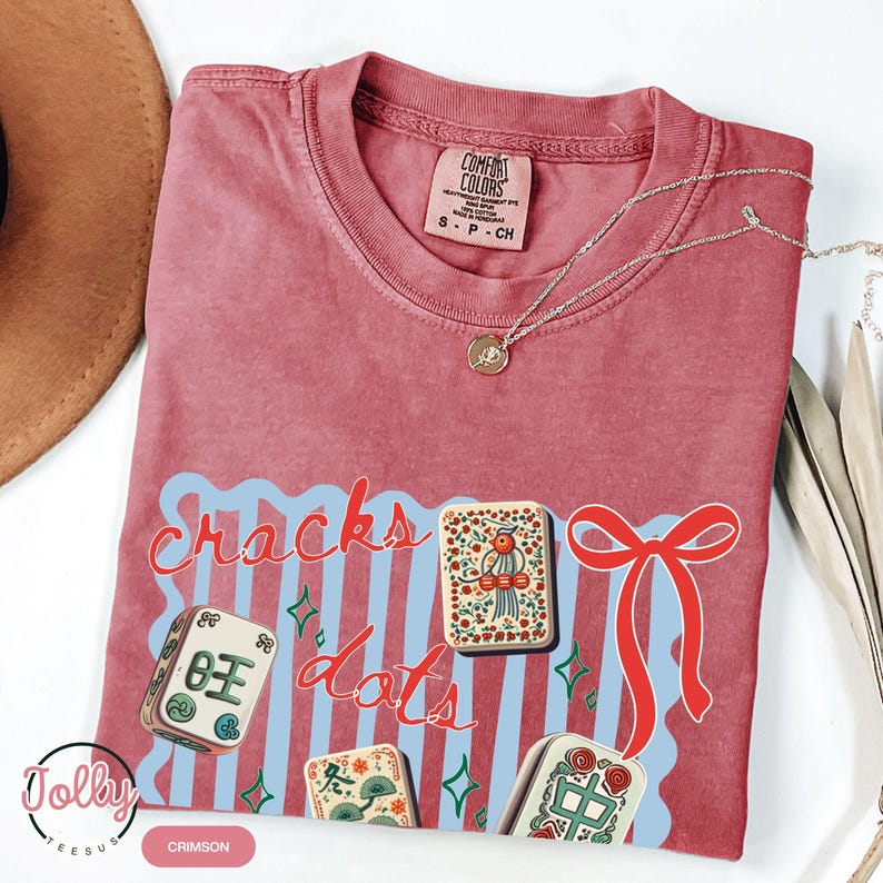 May include: Crimson-colored t-shirt with the words "Cracks Dots" in red script over a light blue wavy design. The shirt features illustrations of Mahjong tiles and a red bow. A gold necklace is draped across the shirt.