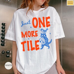 May include: White t-shirt with a graphic design featuring the text "Just ONE MORE TILE" in blue and orange. A blue cat holds a tile, with a bird perched on its head. The shirt has a round neckline and short sleeves.