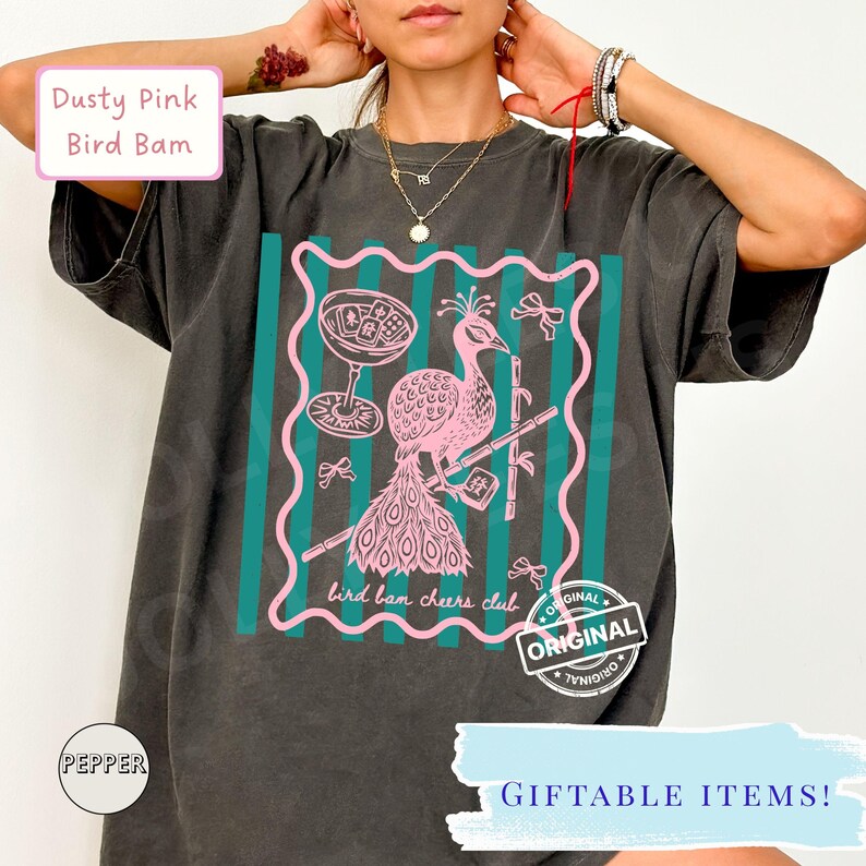 May include: Dark gray t-shirt featuring a pink peacock design with a cocktail glass and bamboo. The text "bird bam cheers club" is included in the design. The shirt also has the text "GIFTABLE ITEMS!" and "ORIGINAL".
