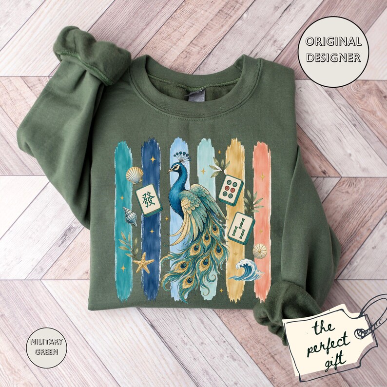 May include: Military green sweatshirt featuring a peacock design with colourful brushstrokes and Asian-inspired imagery. The text "ORIGINAL DESIGNER" is in a circle, and "the perfect gift" is on a tag.