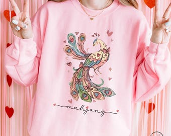 May include: Light pink sweatshirt featuring a colorful peacock design with heart accents and the word "mahjong" in script. The peacock has a bow and the sweatshirt has the text "LIGHT PINK" at the bottom.