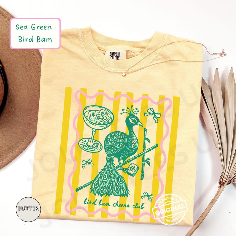 May include: A light yellow t-shirt with a green peacock illustration, a cocktail glass, and bamboo stalks. The design is framed by pink and yellow stripes, with the text "bird bam cheers club". The shirt also has the text "Sea Green Bird Bam".