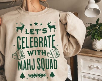 May include: Beige sweatshirt with a green graphic that reads "LET'S CELEBRATE WITH MAHJ SQUAD." The design includes deer, stars, Mahjong tiles, and a festive evergreen border. A comfortable, casual top for game lovers.